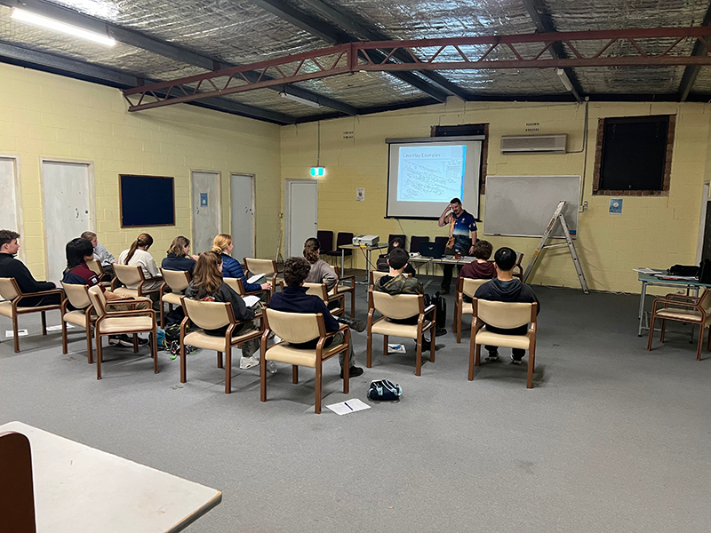 An adult presenting to a group of young people. The presenter is using a projector. There are chairs, a small table and a ladder in the background.