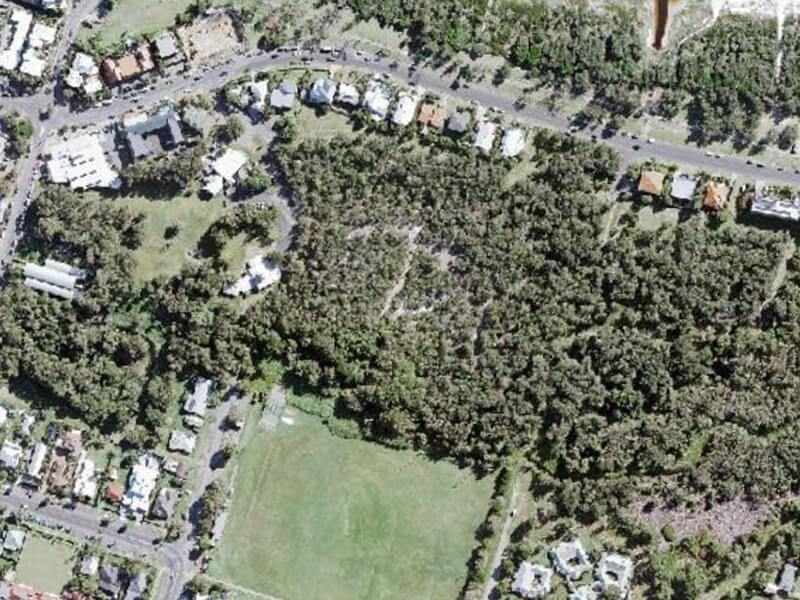 Aerial view of an estate in Byron Bay. In the top of the image, there is a long stretch of buildings facing a main road. Behind the buildings is a large area covered by trees. There are roads on the left and right of the image and buildings dotted along them.